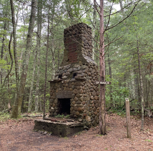 A stone chimney stands in a forest, with a trail sign pointing to "CP Trail" nearby. Lush greenery surrounds the area.