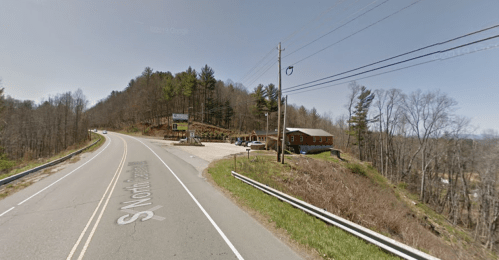 A rural road curves alongside a hillside with a small building and trees in the background on a clear day.