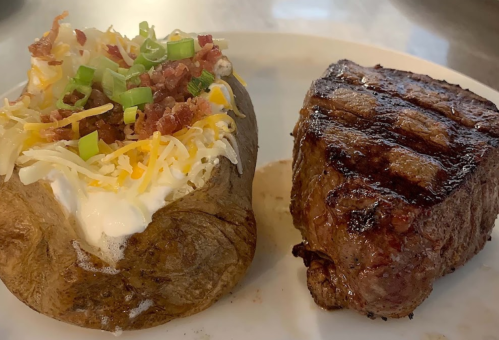 A grilled steak next to a loaded baked potato topped with cheese, bacon, green onions, and sour cream.