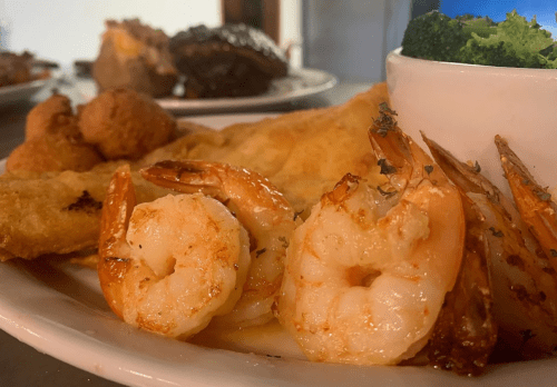 A plate featuring grilled shrimp, fried fish, broccoli, and hushpuppies, showcasing a delicious seafood meal.
