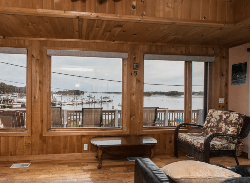 Cozy wooden interior with large windows overlooking a marina and calm water, featuring a comfortable chair and table.