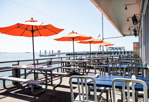 Outdoor dining area by the water, featuring blue tables and orange umbrellas, with a view of a pier in the background.
