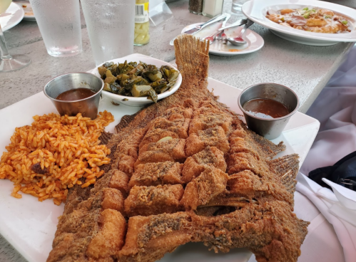 A plate featuring a whole fried fish, rice, greens, and two small bowls of sauce.