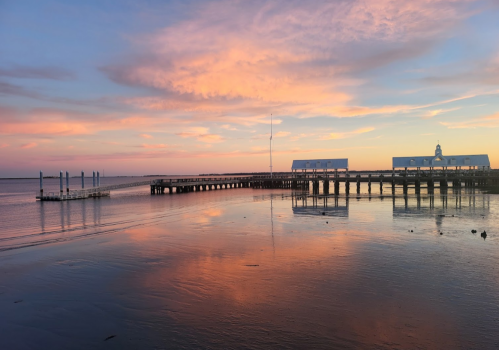 A serene pier at sunset, reflecting vibrant pink and orange hues on calm water.