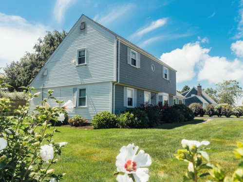 A light blue house with a well-maintained lawn and flowers in the foreground, under a clear blue sky.
