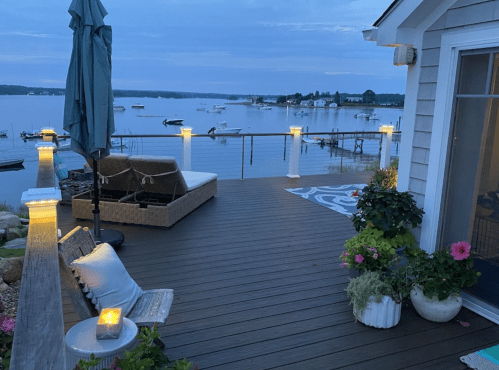 A serene lakeside deck at dusk, featuring a cozy seating area, potted plants, and soft lighting along the railing.