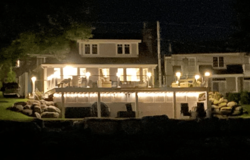 A well-lit house at night with a deck, surrounded by rocks and trees, creating a cozy atmosphere.