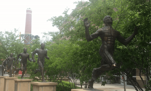 A row of bronze statues depicting runners, surrounded by greenery, with a smokestack in the background.