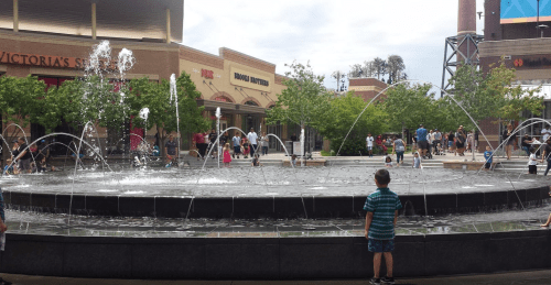 A child stands by a fountain with water jets, surrounded by shoppers and greenery in an outdoor shopping area.