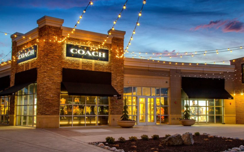 A Coach store exterior at dusk, illuminated by string lights, with large windows and a welcoming entrance.