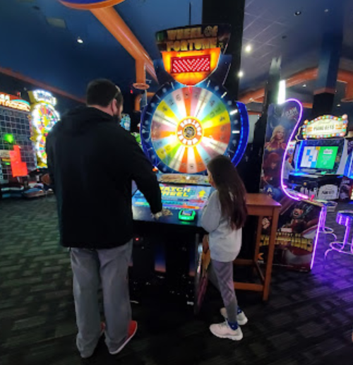 A man and a girl play a colorful arcade game with a spinning wheel in a vibrant game room.