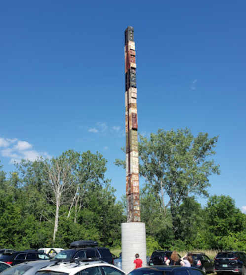 A tall sculpture made of stacked shipping containers, surrounded by trees and parked cars under a clear blue sky.