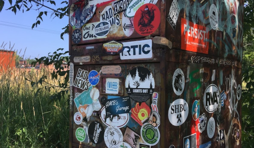A weathered metal box covered in various colorful stickers and graffiti, surrounded by tall grass and trees.