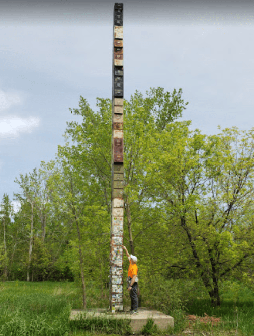 A person in an orange shirt stands beside a tall, colorful totem pole surrounded by green trees and grass.
