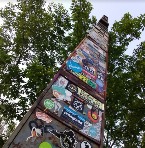 A tall, weathered pole covered in various colorful stickers, surrounded by green trees and a clear sky.
