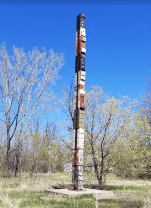 A tall, colorful totem-like structure stands in a grassy area, surrounded by trees under a clear blue sky.