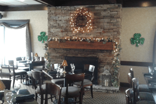 Cozy restaurant interior with a stone fireplace, festive wreath, and green shamrock decorations. Tables set for dining.