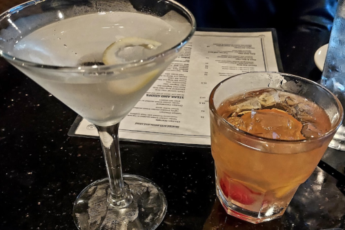 A martini with a lemon slice and a whiskey cocktail with a cherry, set on a dark table with a menu in the background.