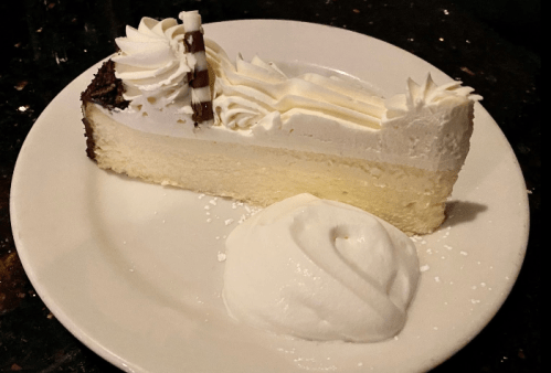 A slice of cheesecake topped with whipped cream and a chocolate garnish, served with a dollop of whipped cream on the side.