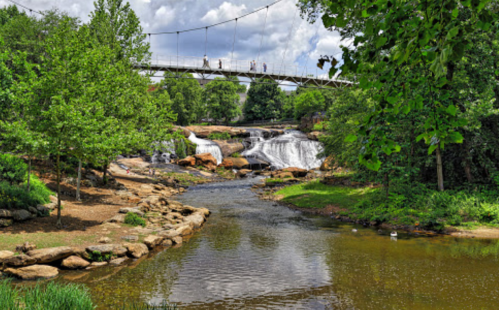 A scenic view of a river with waterfalls, surrounded by greenery and a pedestrian bridge in the background.