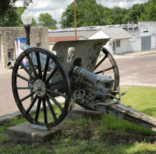 A vintage cannon on a stone pedestal, featuring large wooden wheels and a weathered metal body, set in a grassy area.
