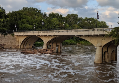 A stone bridge spans a river with swirling water and debris, surrounded by lush greenery and cloudy skies.