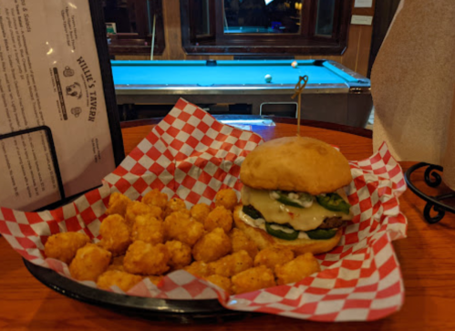 A burger with jalapeños and melted cheese served with a side of tater tots, set on a checkered paper in a tavern.