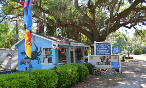 A small seafood market with a blue exterior, flags, and a sign advertising live crabs and fresh shrimp under a large tree.