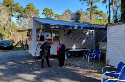 Two people stand outside a food truck in a wooded area, with chairs nearby and a clear blue sky above.