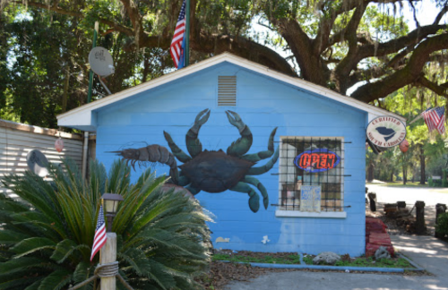 A blue building with a large crab mural, an "Open" sign, and American flags, surrounded by greenery.