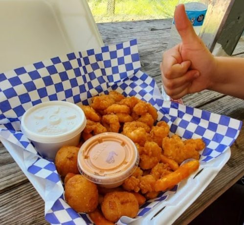 A takeout box filled with fried shrimp, hushpuppies, and dipping sauces, with a hand giving a thumbs up in the background.