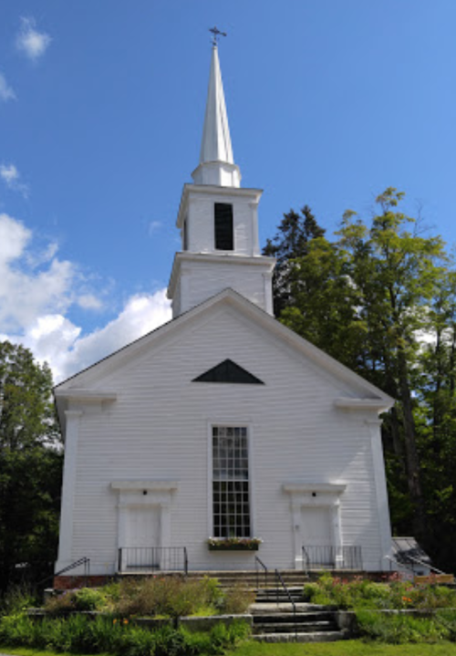 A white church with a tall steeple, surrounded by trees and a blue sky, featuring steps leading to the entrance.
