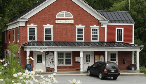 A red brick town hall building with a black roof, featuring a porch and a parked vehicle in front.