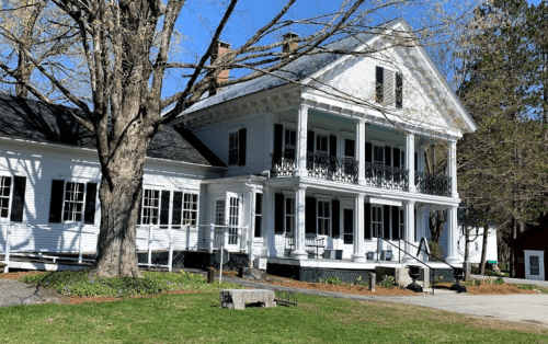 A large white house with a porch, surrounded by trees and a grassy area, under a clear blue sky.