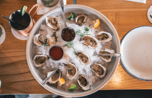 A platter of fresh oysters on ice, garnished with lemon wedges and dipping sauces, served on a wooden table.