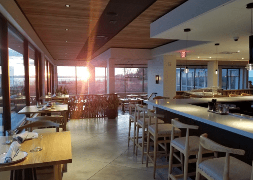 Modern restaurant interior with wooden tables and bar, featuring large windows and a sunset view.