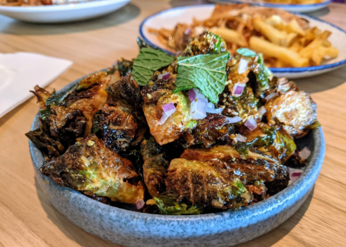 A bowl of crispy Brussels sprouts garnished with mint and diced onions, served on a wooden table.