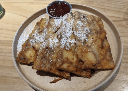 A plate of golden-brown pancakes dusted with powdered sugar, served with a small bowl of red fruit sauce.