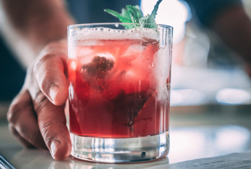 A hand holding a glass of red cocktail with ice and a mint garnish.