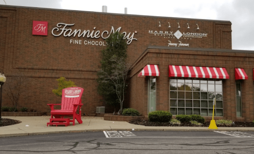 Exterior view of a Fannie May Fine Chocolates store with a red and white striped awning and a large red chair outside.