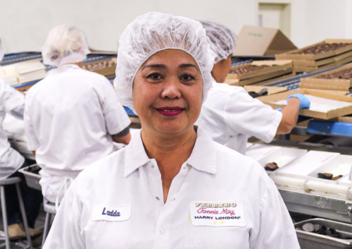 A woman in a hairnet and white uniform stands in a factory, with workers and chocolate trays in the background.