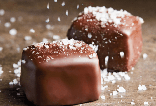 Two chocolate squares topped with a sprinkle of coarse sea salt on a rustic surface.