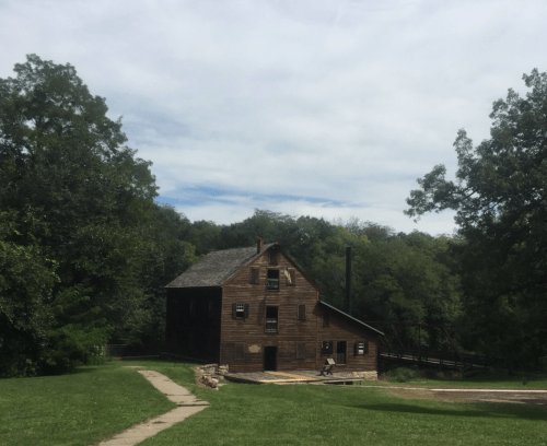 A rustic wooden building surrounded by trees, with a path leading to it under a cloudy sky.