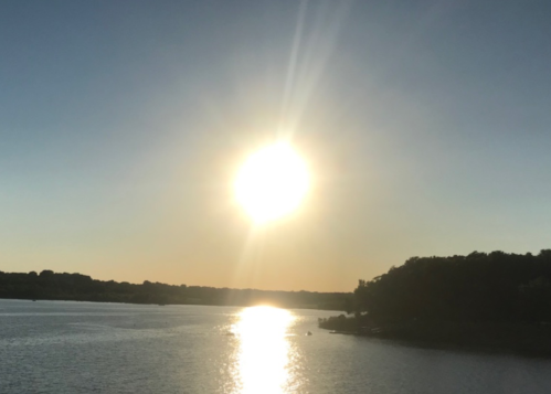 Sunset over a calm lake, with the sun reflecting on the water and silhouettes of trees along the shore.