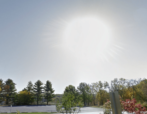 Bright sun shining in a clear sky over a landscape with trees and a parking area.
