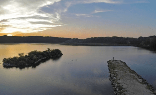 A serene sunset over a calm lake, with a person standing on a rocky pier near a small island.