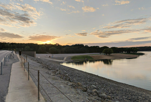A serene lakeside view at sunset, with a path, trees, and calm water reflecting the colorful sky.