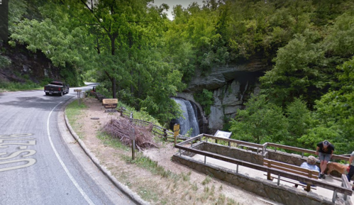 A winding road near a waterfall, with a bench and a person with a dog in a lush green forested area.