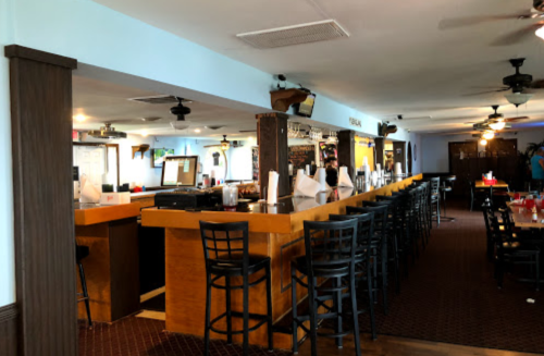 Interior of a bar with a long wooden counter, high stools, and a casual dining area. Brightly lit and inviting atmosphere.
