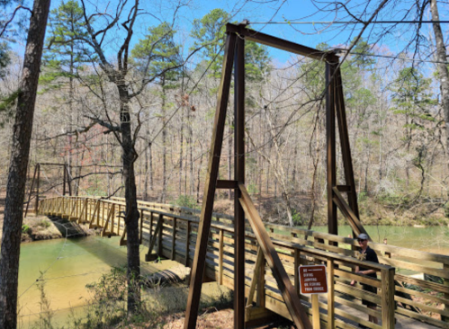A wooden suspension bridge spans a calm river, surrounded by trees and a clear blue sky.